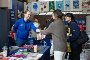 一个穿着蓝色U / R四分之一拉链卫衣的人坐在健康博览会上，微笑着向两个学生打招呼，并与他们握手. (繁荣节2024)